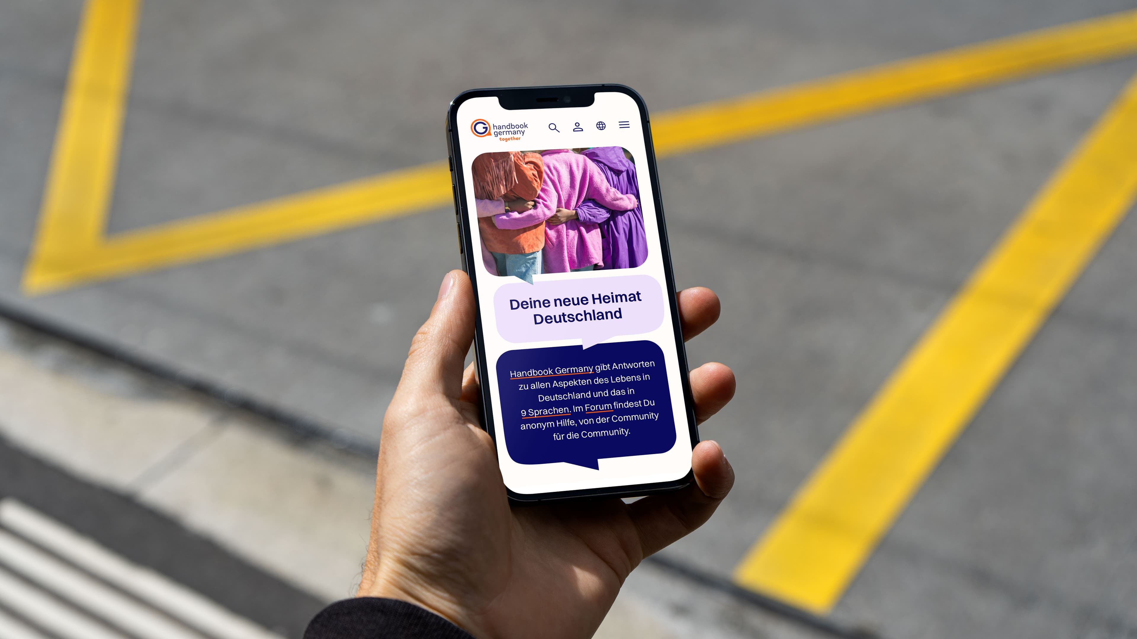 Hand hält ein Smartphone mit der Website „Handbook Germany“ vor einem grauen Boden mit gelben Linien.