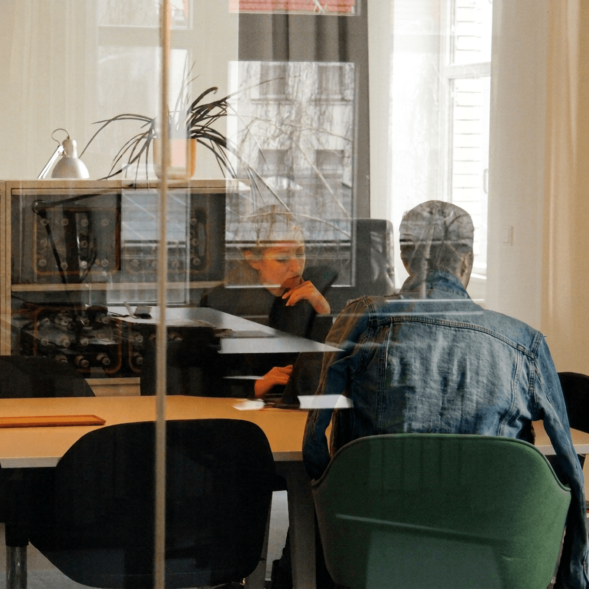 Zwei Personen sitzen sich an einem Tisch in einem hellen Raum gegenüber, aufgenommen durch eine Glasscheibe mit Spiegelungen.