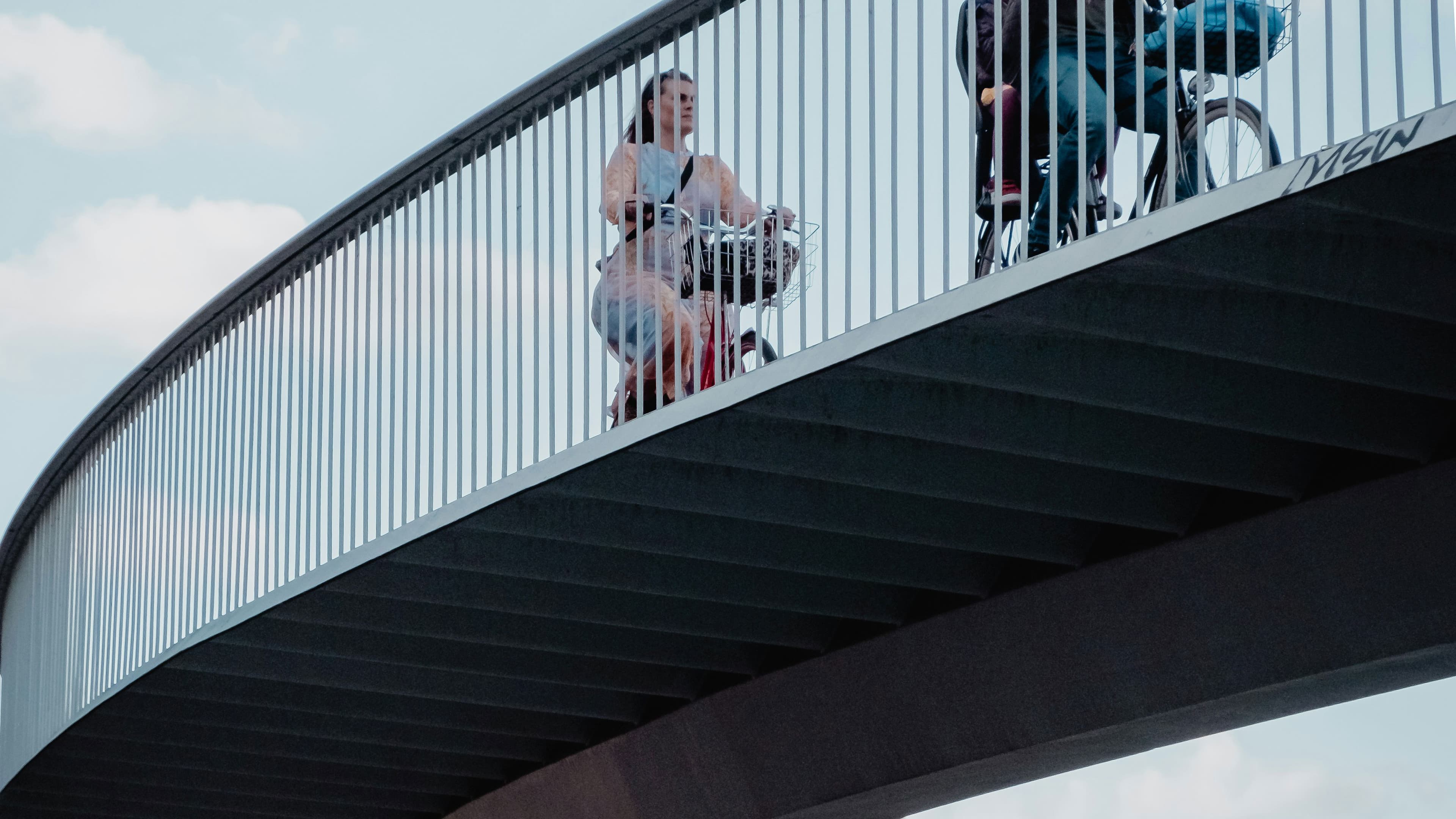 Two people on bicycles on a bridge with a railing, photographed from below.